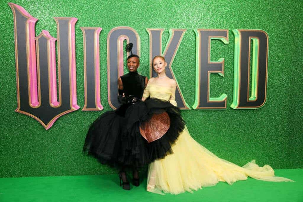 Movie actresses Cynthia Erivo in a black dress and Ariana Grande in a cream colored dress attend the European Premiere of the movie "Wicked: Part One" at The Royal Festival Hall. They pose in front of a green background with the Wicked logo in black lettering.